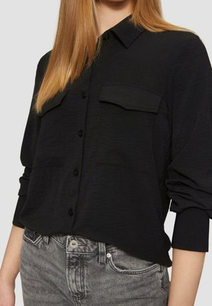 Femme portant une chemise noire boutonnée avec poches poitrine et manches retroussées, associée à un jean gris délavé.