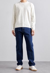Personne portant un pull texturé blanc à manches longues, un jean bleu droit et des baskets blanches, debout sur un sol gris contre un mur uni.