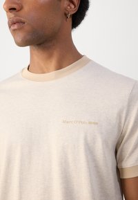 Beige tričko s kruhovým výstřihem a kontrastním světle hnědým žebrovaným límečkem, na hrudi s jemným zlatým textem "Marc O'Polo DENIM".