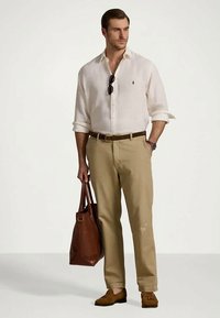 Mężczyzna w podwiniętej białej koszuli z guzikami i beżowych spodniach, trzymający brązową skórzaną torbę, z okularami przeciwsłonecznymi zawieszonymi na kołnierzu koszuli.