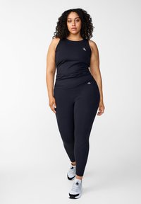 Schwarzes sportliches Tanktop und Leggings aus elastischem Material, mit einem figurbetonten Design und einem kleinen Logo auf dem Top. Abgerundet mit weißen Sneakern.