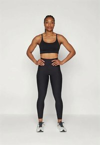 Atleetinainen seisoo itsevarmasti kädet lanteilla, pukeutuneena mustaan urheiluliiviiin, korkeavyötäröisiin legginsseihin ja valkoisiin juoksukenkiin tasaisen taustan edessä.