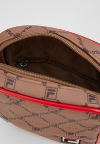 Sac en tissu marron avec motif en relief du logo FILA, accent de zip rouge et poche zippée intérieure, présentant une texture lisse et une forme arrondie.