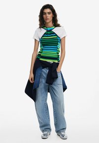 Top a righe, aderente, verde, blu e nero con una manica bianca, abbinato a jeans larghi di un blu chiaro e a un maglione blu navy annodato in vita.