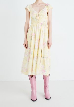 Woman wearing a yellow floral midi dress with short sleeves and pink cowboy boots, standing against a plain white background.