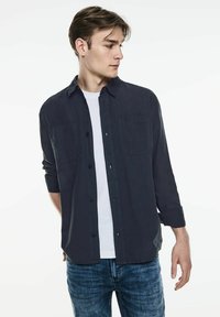 Marinblå button-up skjorta, bomullstyg, två bröstfickor, långa ärmar uppvikta, bärs över en vit t-shirt, kombinerat med blå jeans.