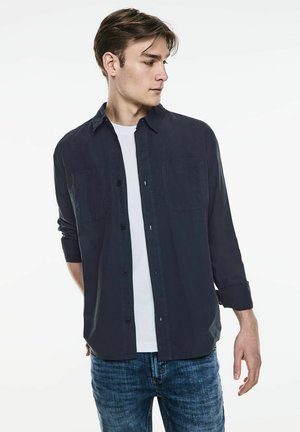 Camisa azul marino de botones, tejido de algodón, con dos bolsillos en el pecho, mangas largas arremangadas, llevada sobre una camiseta blanca, combinada con jeans azules.