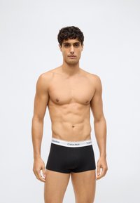 En mann uten skjorte med kort, mørkt hår iført svarte Calvin Klein boksershorts som står mot en enkel, lys bakgrunn.
