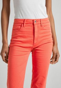 Vaqueros de coral brillante con un diseño ajustado, que presentan una cintura alta, estilo de cinco bolsillos y detalles de costura sutiles. Textura de mezclilla suave.