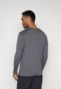 Langermet grå skjorte laget av glatt stoff med rund hals. Har en liten oransje etikett nær kanten, enkelt design og tettsittende ermer.