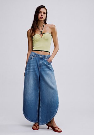 Femme portant un haut bustier jaune à col licou, un jean bleu large à ourlets arrondis et des sandales rouges à talons, debout les mains dans les poches.