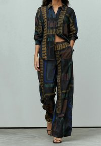 Donna che cammina indossando una camicia nera ampia e decorata con un motivo a forme astratte colorate, abbinata a pantaloni larghi dello stesso stile, con tacchi aperti neri.