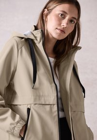 Veste à capuche beige clair avec fermeture éclair frontale, poignets élastiques et cordons de serrage noirs. Présente une texture lisse et mate ainsi que des poches latérales.