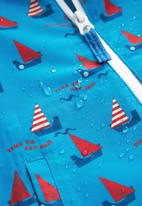 Tissu imperméable bleu avec des motifs de voiliers rouges et blancs, des gouttes d'eau et une fermeture éclair blanche avec une tirette en tissu bleu.