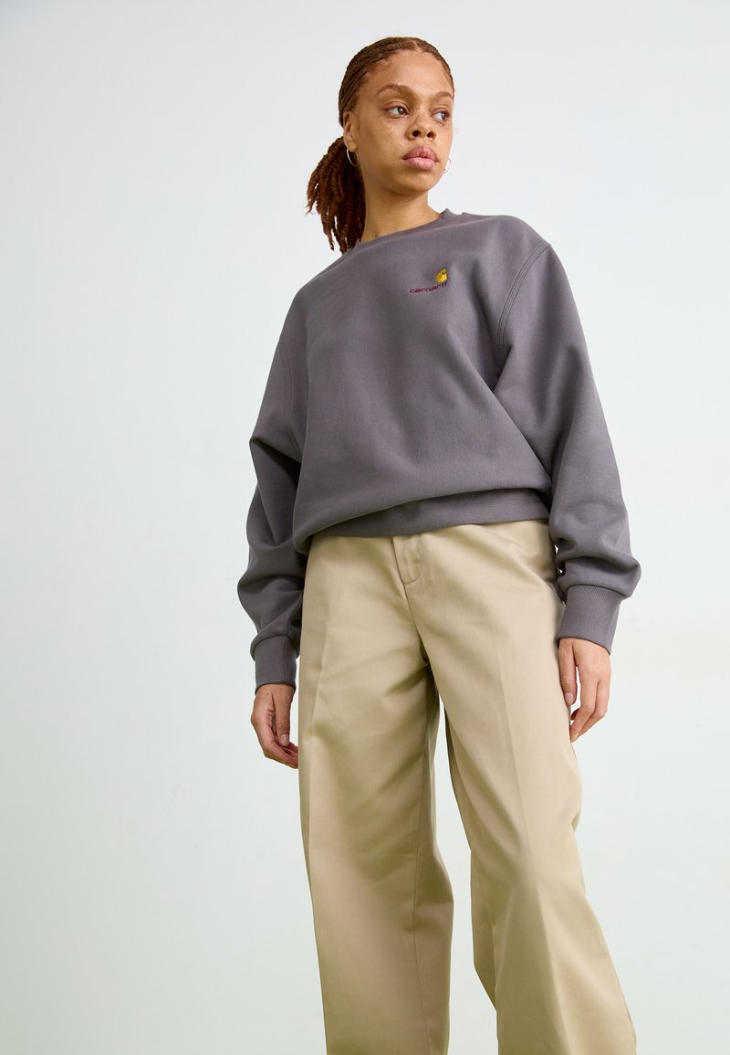 Grå sweatshirt med ribbad mudd och halsringning, med en liten broderad logotyp. Kombinerad med ljusbeiga vida byxor.