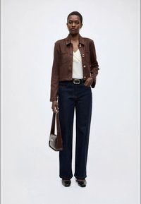 Braune Wildlederjacke mit goldenen Knöpfen, cremefarbenes Oberteil, dunkelblaue Jeans mit weitem Bein und schwarze Schuhe. Modell hält eine braune Handtasche. Einfarbiger Hintergrund.