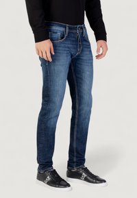 Jeans in denim blu scuro con vestibilità slim, caratterizzati da cuciture a contrasto, design a cinque tasche e chiusura con zip. Abbinati a sneakers nere.