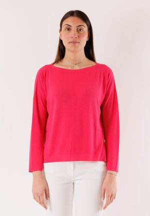 Donna in piedi, rivolta verso la fotocamera, che indossa una maglia a maniche lunghe di un rosa acceso e pantaloni bianchi, su uno sfondo chiaro e uniforme.