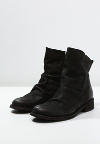Par de botas de cuero negro hasta el tobillo con superficie texturizada, cañas caídas y tacones bajos apilados sobre un fondo blanco.