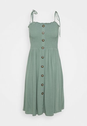 Saliegroene mouwloze jurk met geplooide corsage, schouderbandjes met strik en een afsluiting met bruine knopen aan de voorkant.