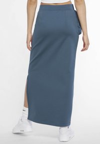 Jupe longue bleue ajustée en jersey avec une fente latérale, présentant une texture lisse et une taille cintrée, assortie à des baskets blanches.