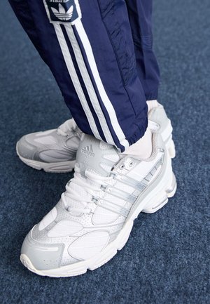 Weiße und silberne Adidas-Sneaker, getragen mit weißen Socken und dunkelblauen Adidas-Trainingshosen mit weißen Streifen auf blauem Teppich.