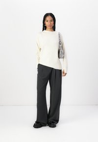 Pull en maille côtelée beige clair, coupe ample, avec de longues manches ; associé à un pantalon large gris foncé. Le modèle porte un sac à épaule gris.