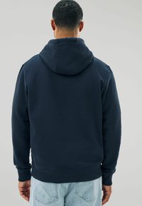 Sudadera oscura en color marino con bolsillo canguro, dobladillo y puños acanalados, y capucha con cordón. Textura suave, diseño casual y corte relajado.