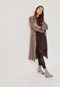 Długi brązowy płaszcz z mieszanki wełny z wycięciami, bocznymi kieszeniami i srebrnymi guzikami. Noszony na brązową sukienkę, uzupełniony szarymi zamszowymi botkami za kostkę.