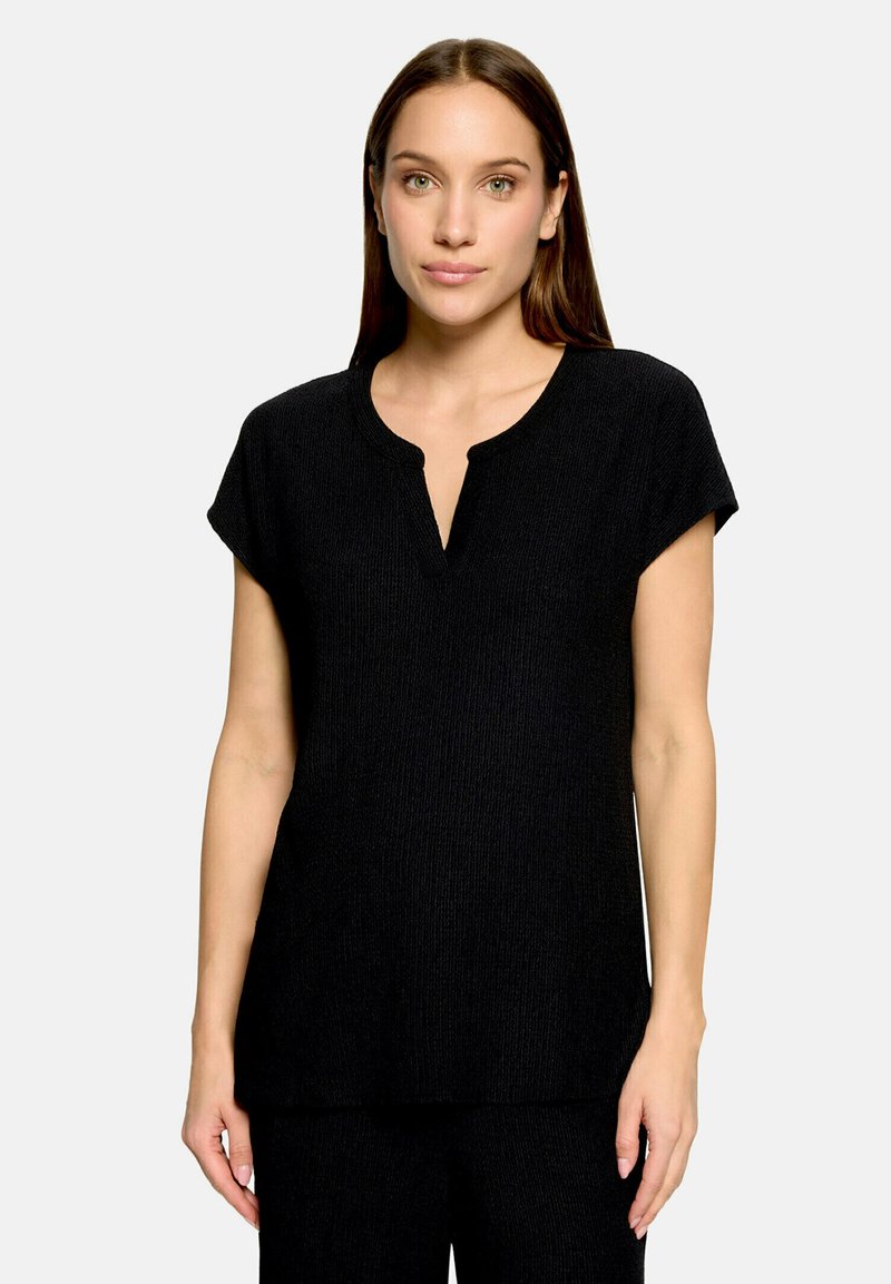 Femme aux longs cheveux bruns portant un haut en maille noir à manches courtes avec un col rond et une petite fente à l'avant, debout sur un fond uni.