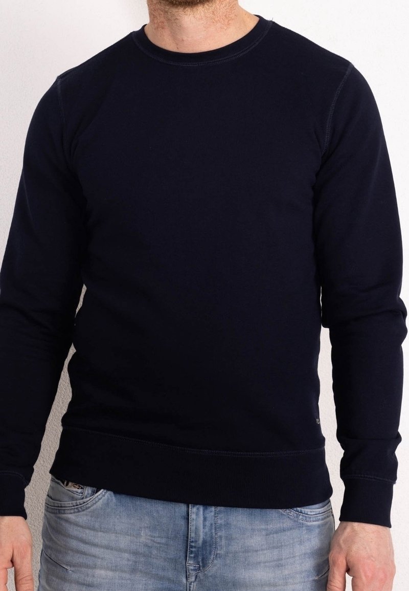 Homme portant un pull noir uni à manches longues et col rond ainsi qu'un jean bleu clair, debout devant un fond uni clair.