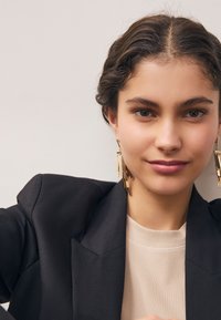 Guldstatementörhängen med lager på lager av geometriska former; modell som bär en svart blazer och en krämfärgad ribbad topp, slät hud, naturlig makeup.