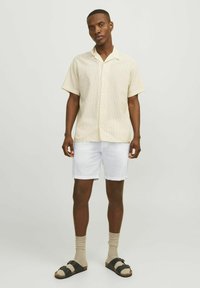 Camisa de botones de manga corta en un ligero color amarillo con un sutil patrón texturizado, combinada con pantalones cortos blancos y sandalias negras con calcetines beige.