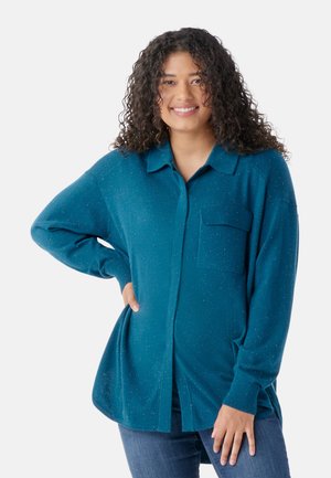 Chemise à manches longues teal en tissu tricot doux, avec un col, une poche avant et un détail légèrement chiné. Coupe décontractée avec fentes sur les côtés.