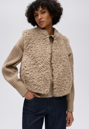 Donna con capelli corti e ricci che indossa un gilet beige dalla texture riccia sopra un maglione a coste con cerniera e pantaloni blu scuro su uno sfondo semplice.