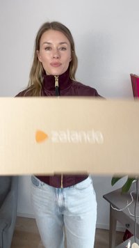 Brun pappkartong med Zalando-logotyp hålls framför en person som bär en mörk burgunderjacka och ljusblå jeans mot en enkel vägg.