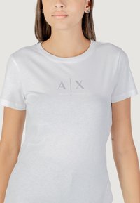 Bijela pamučna majica s okruglim izrezom, kratkim rukavima i srebrnim logotipom "A|X ARMANI EXCHANGE" otisnutim na sredini prednje strane.