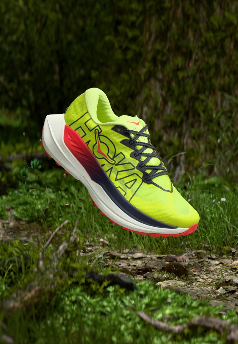 Φωτεινό κίτρινο και κόκκινο παπούτσι για τρέξιμο σε μονοπάτι με μαύρα κορδόνια, στον αέρα πάνω από δάσος με γρασίδι και μικρές πέτρες.