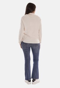 Top lungo beige chiaro a maniche lunghe con colletto drappeggiato, abbinato a jeans a zampa d'elefante blu e sneakers bianche, che mette in mostra un look casual visto da dietro.