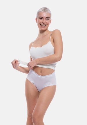 Giovane donna con capelli biondi corti che sorride, indossando un canottiera bianca e biancheria intima alta con pizzo, sollevando leggermente la maglietta.