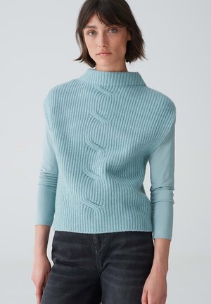 Hellblauer Strickpullover mit hohem Kragen und gerippter Textur, der ein geflochtenes Kabelmuster in der Mitte aufweist. Wird über einem Langarmshirt getragen.