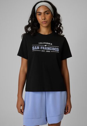 Kobieta o czarnych kręconych włosach, nosząca szarą opaskę, czarną koszulkę z napisem "San Francisco California" oraz jasnobłękitne szorty na szarym tle.
