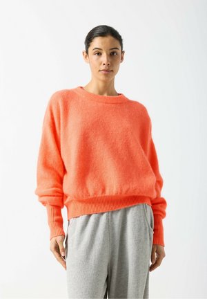 Helles oranges, flauschiges Sweatshirt mit Rundhalsausschnitt und geripptem Saum, kombiniert mit hellgrauen Freizeithosen. Weiche Textur und lockere Passform.