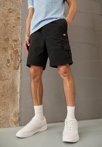 Schwarze Cargo-Shorts aus Baumwolle mit einer Seitentasche und Logo-Patch, kombiniert mit weißen Sneakern und gerippten weißen Socken.