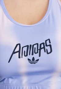 Svetlo vijoličasta športna majica brez rokavov z velikim črnim logotipom "Adidas" in emblemom trefoil. Gladek material z zaobljenim ovratnikom.