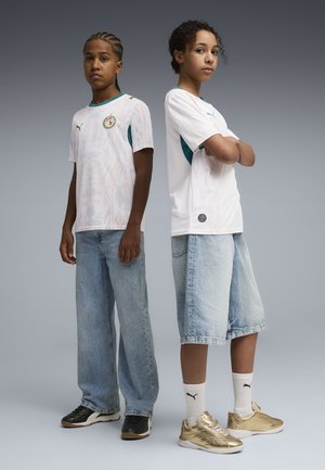 Dos jóvenes están de espaldas, vistiendo camisetas blancas de fútbol de Senegal, pantalones de mezclilla y zapatillas Puma, contra un fondo gris.