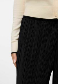 Svarta veckade byxor med en slät midjeband, kombinerat med en ljusbeige tröja med knappstängning och kontrasterande svarta detaljer.