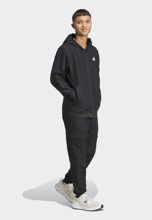 Svart träningsoverall med huva och dragkedja fram, i kombination med matchande cargobyxor. Vitt logotyp på bröstet, kompletterad med beige sneakers.