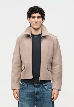 Jeune homme aux cheveux bouclés et foncés portant une veste zippée marron clair sur une chemise noire et un jean bleu, debout devant un fond clair uni.