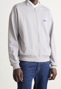 Mand iført en lys grå zip-up bomberjakke med et lille blåt "HUGO" logo og blå jeans, der står op ad en ensfarvet baggrund.